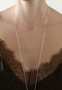 Silverkedja hänger över ett mörkt tyg med intrikat spetskant som har ett blommönster och fransiga kanter.