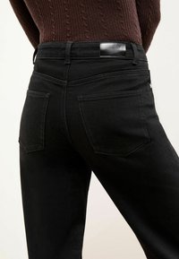 Vaqueros de denim negro con un diseño ajustado, que cuentan con dos bolsillos traseros y un parche de cuero suave en la cinturilla. Textura suave y costura sencilla.