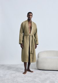 Accappatoio verde oliva con trama a nido d'ape, cintura annodata in vita e tasche laterali, caratterizzato da un marchio discreto. Il modello è in piedi a piedi nudi su un tappeto grigio.