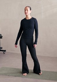 Maglietta nera a maniche lunghe e pantaloni alla flare attillati realizzati in materiale elastico. In piedi su un tappeto verde contro un muro neutro.