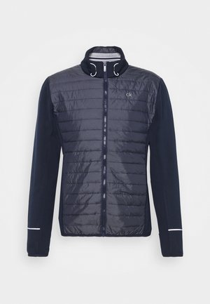 Laivastonsininen takki, jossa tikattu etuosa, sileät hihat ja korkea kaulus. Täysipitkä vetoketju ja hillityt heijastavat yksityiskohdat hihansuissa.