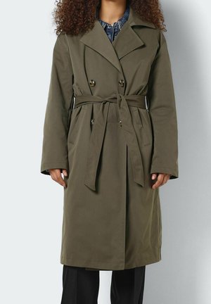 Kvinde iført olivengrøn dobbeltbrystet trenchcoat med bælte bundet i taljen, over mørke bukser og denimskjorte.