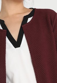 Detailní záběr na osobu, která má na sobě bílou blůzu s černým lemem pod texturovaným, tmavě červeným kardiganem s otevřeným předním dílem.