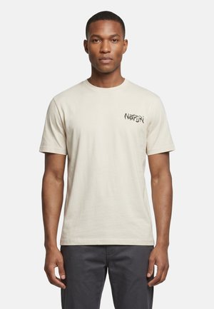 Man die een effen lichtbeige T-shirt met korte mouwen draagt met een zwart tekstlogo op de linkerborst, gecombineerd met een donkere broek, staand tegen een witte achtergrond.