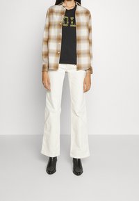 Ruutupaita ruskean ja kermaisen sävyssä mustan t-paidan päällä, jossa on keltainen graafinen kuvio. Yhdistetty valkoisiin leveälahkeisiin farkkuihin ja mustiin nilkkureihin.