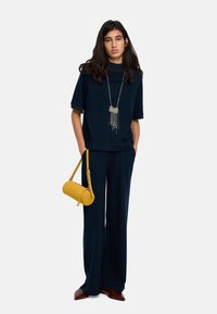 Granatowa bluzka z krótkim rękawem i szerokie spodnie, uzupełnione srebrnym naszyjnikiem oraz żółtą zamszową torebką na ramię. Gładkie tekstury i luźny krój.