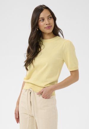 Femme aux longs cheveux bruns portant un pull à manches courtes jaune clair et un pantalon beige à cordon de serrage, debout avec une main dans la poche.