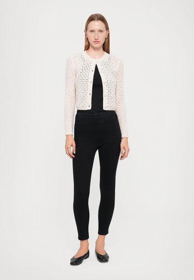 Neulejakku vaaleanpunaisena, jossa on avoin kuvio, pyöreä pääntie ja nappikiinnitys, yhdistettynä mustiin slim-fit legginsseihin ja ballerinoihin.