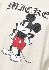 Camiseta blanca con un estampado desgastado de Mickey Mouse, en colores negro y rojo, con orejas redondas y guantes blancos, brazos en las caderas.
