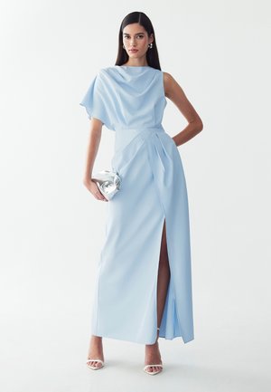 Mujer con un vestido asimétrico azul claro con una apertura alta, llevando un bolso de mano plateado y tacones blancos abiertos en los dedos.