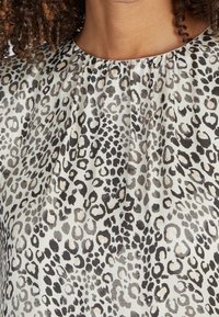 Blouse à imprimé léopard avec une base blanche ornée de taches noires et grises, un plissage subtil autour de l'encolure, et une texture de tissu lisse.