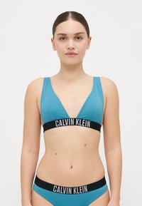 Ljusblå bikiniuppsättning med ett triangulärt topp med ett svart "CALVIN KLEIN"-band, matchande bottnar och ett mjukt, stretchigt tyg.