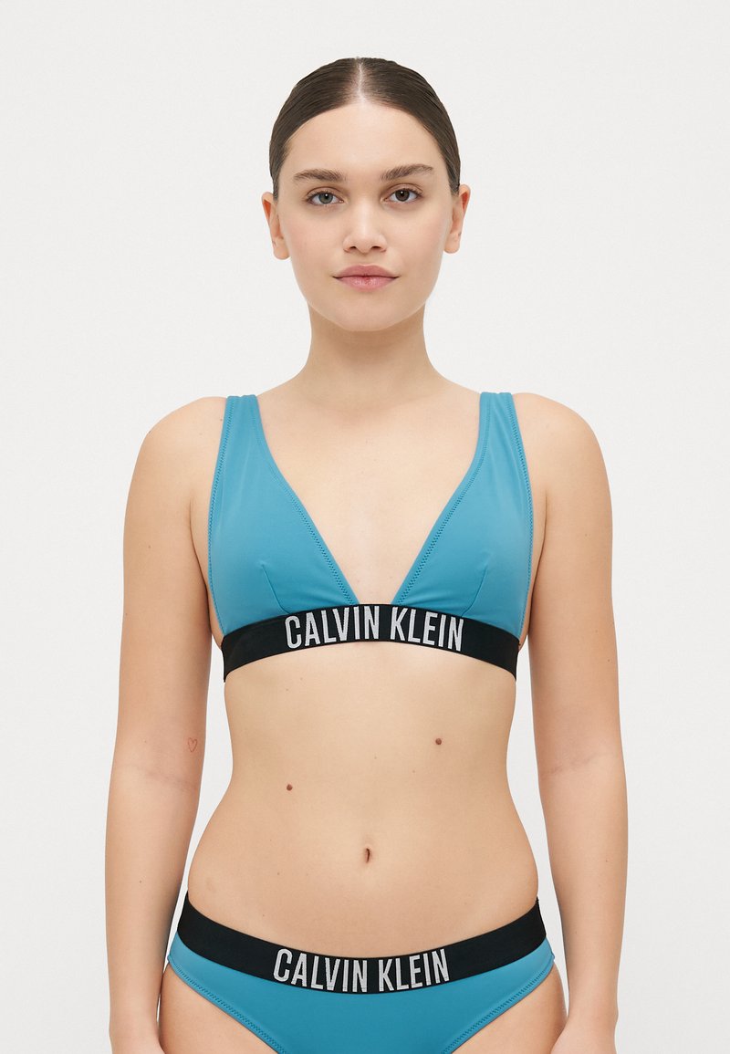 Ljusblå bikiniuppsättning med ett triangulärt topp med ett svart "CALVIN KLEIN"-band, matchande bottnar och ett mjukt, stretchigt tyg.