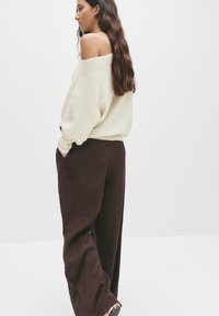 Cremfarbener Off-Shoulder-Pullover mit lockerer Passform, gerippten Bündchen und braunen weiten Hosen. Das Outfit zeichnet sich durch ein minimalistisches, strukturiertes Erscheinungsbild aus.
