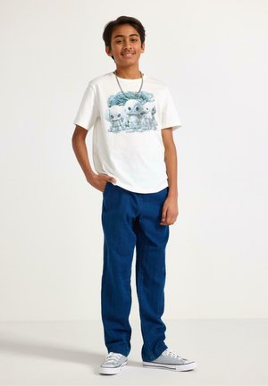 Teenagedreng smilende iført hvid grafisk t-shirt med tre tegneserie-robotter, blå jeans, grå sneakers og en sølvkæde.