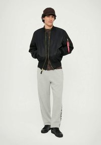Black bomber jacket with zipper, ribbed cuffs and hem, red tag detail. Worn over a brown shirt, paired with grey sweatpants and black shoes.