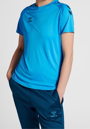 Person trägt blaue hummel-Sportbekleidung mit Händen in den Taschen einer marineblauen hummel-Trainingshose vor einfarbigem Hintergrund.