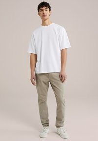 Witte t-shirt met korte mouwen van gladde stof, gecombineerd met lichtbeige broek. Model staat voor een neutrale achtergrond en draagt witte sneakers.
