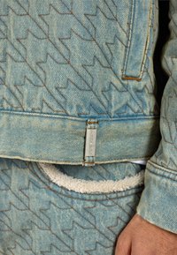 Τζάκετ από τζην σε ανοιχτό μπλε χρώμα με υφή ζιγκ-ζαγκ και καφέ ραφές. Διαθέτει ετικέτα στο τελείωμα και τσέπη επενδεδυμένη με φλις.