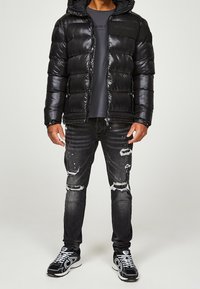Sort pufferjakke med hætte, blank tekstur, lynlås lukning og logo detalje; parret med slidte grå jeans og sorte sneakers.
