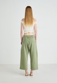 Kvinde med langt blondt hår iført en off-shoulder hvid crop top, brede grønne bukser og hvide højhælede sandaler, stående med ryggen til.