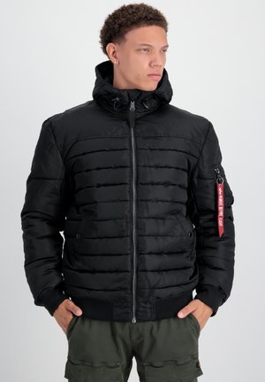 Zwarte pufferjack met ritssluiting, hoge kraag en geribde manchetten. Heeft zijzakken en een rode stoffen label op de mouw. Glad textuur.