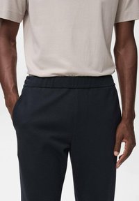 Marine stretchbroek met een platte tailleband en zijzakken. Gеdragen met een lichtbeige T-shirt met korte mouwen. De stof heeft een gladde textuur.