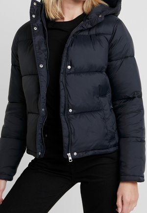 Personne portant une doudoune bleu marine foncé avec fermeture éclair et boutons-pression, pantalon noir et haut noir sur un fond clair uni.