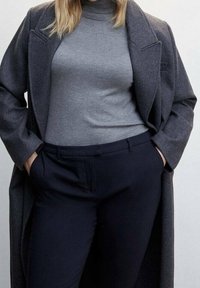 Manteau gris surdimensionné porté sur un pull à col roulé gris, associé à un pantalon bleu marine foncé. Tissu texturé avec coutures visibles et poches.