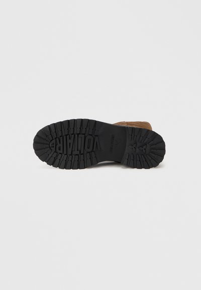 Καφέ σουέντ μποτάκι με υφή μαύρης καουτσούκ σόλας, που διαθέτει βαθιά ανάγλυφα και ένα λογότυπο. Η φτέρνα είναι ελαφρώς ανυψωμένη, αυξάνοντας τη σταθερότητα.