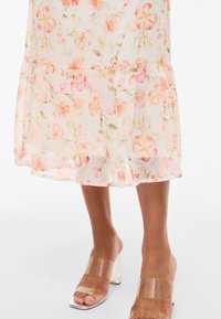 Lenge, áttetsző midi ruha rózsaszín és narancssárga virágmintával, áttetsző pántos magas sarkú szandállal, mezítláb viselve.