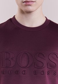 Camiseta de algodón burdeos de cuello redondo con letras en relieve "BOSS" y "HUGO BOSS" en la parte delantera, con una superficie de tejido texturizada.