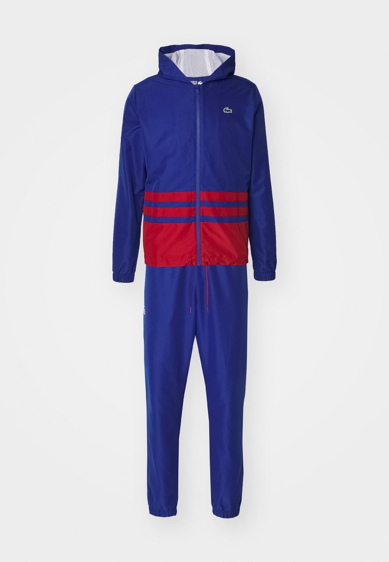 Giubbotto a vento blu con una sezione a strisce rosse in vita, dotato di cappuccio e chiusura con zip. Pantaloni blu coordinati con vita elastica.