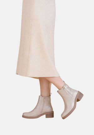 Bottines argentées métalliques avec une finition texturée, zips latéraux et talons empilés marron, portées avec une jupe beige mi-mollet.