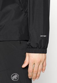 Gros plan d'une main portant une veste noire avec un poignet élastique marqué "DRY VENT" et un pantalon noir avec un logo mammouth.