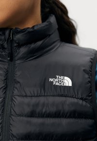 Crna izolirana prsluk s rombi teksturom, visokim ovratnikom i prednjim zatvaračem. Na prsima se nalazi bijeli logo "The North Face".