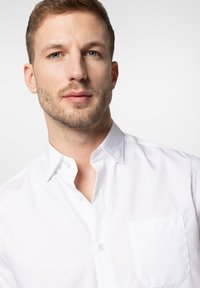 Biała koszula z guzikami, z kieszenią na piersi, klasycznym kołnierzykiem i subtelnymi detalami przeszycia. Gładka faktura materiału, minimalistyczny design.