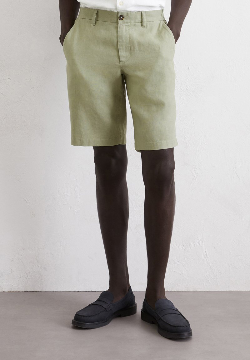 Light green linen shorts with a straight cut, featuring front pockets and a single button closure. Worn with dark loafers.