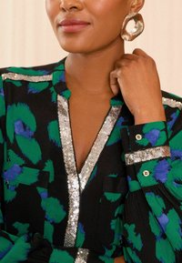 Fekete blúz zöld és kék absztrakt mintával, ezüst flitter díszítéssel a galléron és az ujjakon, valamint két elülső zsebbel.