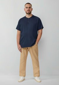 Granatowa koszulka henley z krótkim rękawem i trzema guzikami, zestawiona z beżowymi spodniami i białymi trampkami. Materiał wydaje się gładki i casualowy.