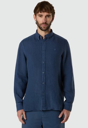 Uomo in piedi, rivolto verso avanti, che indossa una camicia blu navy a maniche lunghe con bottoni e un piccolo logo sul petto.