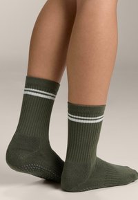 Olívazöld pamutkeverék zokni bordázott textúrával, a szegélyén két fehér csíkkal és mintás csúszásgátló talppal.