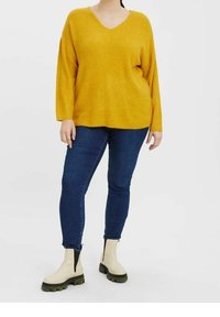 Maglione giallo senape lavorato a maglia con scollo a V, abbinato a jeans blu scuro e stivaletti color crema con suola verde.