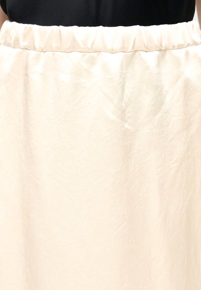 Φούστα από εκρού υλικό με πιέτες, εμφανιζόμενη από τη μέση μέχρι το μέσο του μηρού, φορεμένη πάνω από μαύρο τοπ.