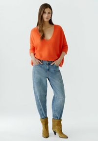 Oranger, übergroßer Strickpullover mit tiefem V-Ausschnitt, kombiniert mit hellblauen, hoch taillierten Jeans und khakifarbenen, hohen Stiefeletten.