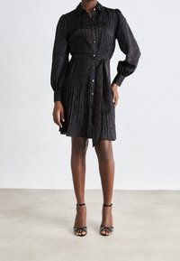 Vestido negro de botones con textura plisada, mangas largas y detalle de cintura ceñida con cinturón. Combinado con tacones negros con tiras.