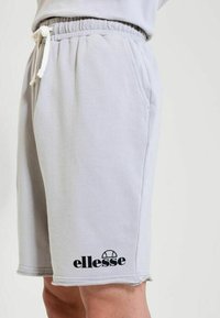 Pantalones cortos ellesse gris claro con cintura elástica y cordón blanco en la parte inferior del torso y la parte superior de las piernas de una persona.