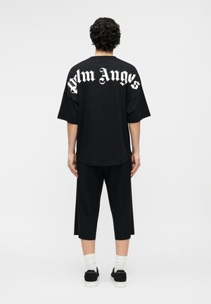 Άτομο με κοντά σγουρά μαλλιά, ντυμένο με άνετο μαύρο πουκάμισο με την επιγραφή «Palm Angels» και μαύρο κοντό παντελόνι, στέκεται γυρισμένο με την πλάτη σε λευκό φόντο.