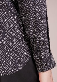 Camicia nera con bottone davanti, decorata con un motivo geometrico a diamante bianco e un sottile dettaglio a forma di teschio sul polsino, realizzata in tessuto leggero.