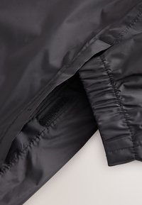 Manche de veste noire avec poignet élastique et fermeture Velcro visible sur la couture intérieure.